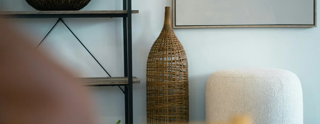 Detail of woven wicker vase, upholstered stool and dark metal shelf against a pale wall